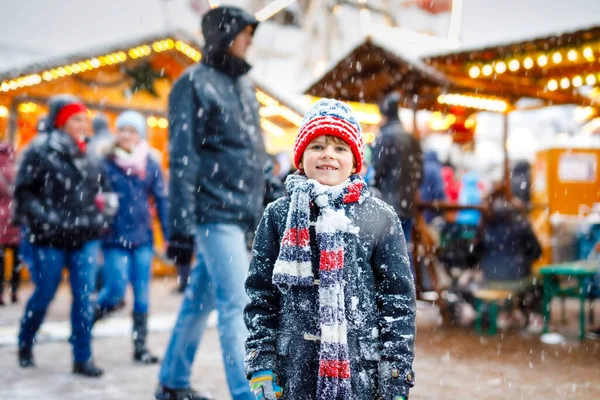 Pequeño niño lindo que se divierte en el mercado tradicional alemán de Navidad durante las fuertes nevadas.. Niño feliz disfrutando del mercado familiar tradicional en Alemania, Munich. Chico risueño en ropa colorida — Foto de Stock