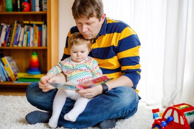 Genç baba, sevimli küçük kızıyla kitap okuyor. Gülümseyen güzel bir çocuk ve erkek evdeki oturma odasında birlikte oturuyorlar. Yeni yürümeye başlayan çocuk babaya kulak veriyor.