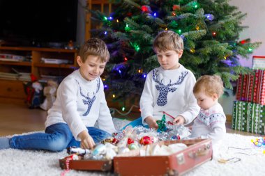 İki küçük oğlan ve sevimli kız bebek Noel ağacını eski model oyuncaklar ve toplarla süslüyorlar. Aile tatili için aile hazırlık kutlaması. Evde üç çocuk, erkek ve kız kardeş var..