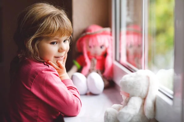 Schattig peuter meisje zit bij het raam en kijkt uit op regenachtige dag. Dromend kind met pop en knuffel die zich gelukkig voelen. Zelfisolatieconcept tijdens de corona-viruspandemie. Eenzame jongen. — Stockfoto