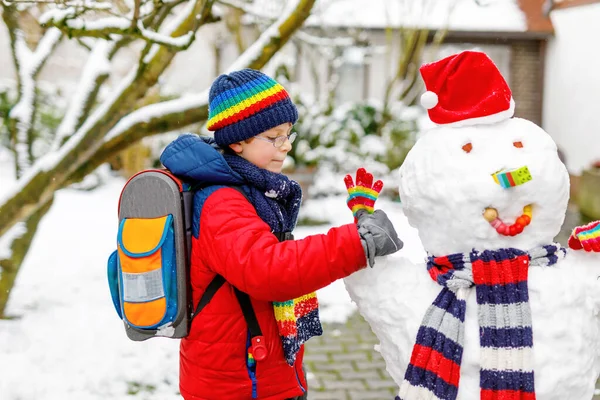 Kleiner Schuljunge in bunten Klamotten, mit Brille und Rucksack, der nach dem Ende der Grundschule Spaß mit dem Schneemann hat — Stockfoto