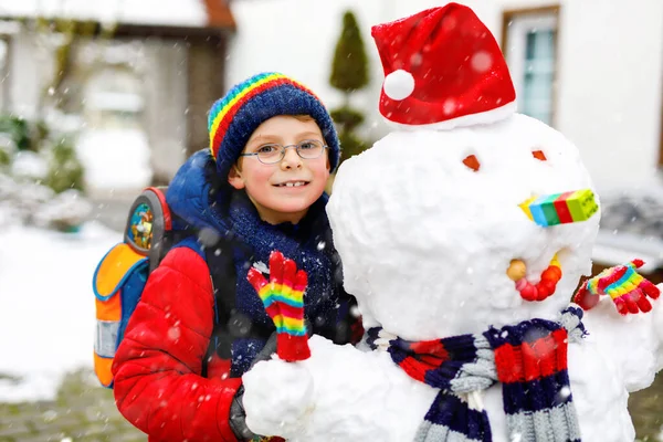 Kleiner Schuljunge in bunten Klamotten, mit Brille und Rucksack, der nach dem Ende der Grundschule Spaß mit dem Schneemann hat — Stockfoto