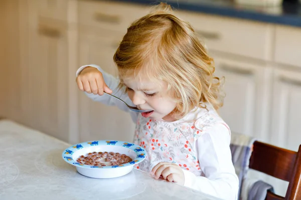 Menina adorável criança comendo cereais saudáveis com leite para o café da manhã. Bonito bebê feliz criança em roupas coloridas sentado na cozinha e se divertindo com a preparação de aveia, cereais. Interior em casa — Fotografia de Stock