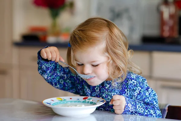 おいしい幼児の女の子は朝食のためにミルクと健康的なシリアルを食べています。キッチンに座って、オート、シリアルを準備するのが楽しいカラフルな服のかわいい幸せな赤ちゃん。家の中で — ストック写真