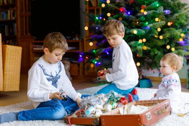 İki küçük oğlan ve sevimli kız bebek Noel ağacını eski model oyuncaklar ve toplarla süslüyorlar. Aile tatili için aile hazırlık kutlaması. Evde üç çocuk, erkek ve kız kardeş var..