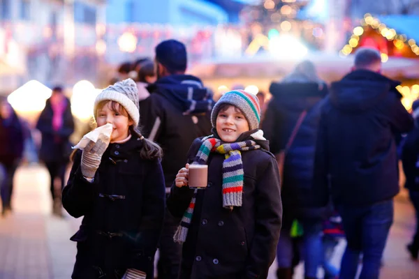 Cute little kids girl and boy having fun on traditional Christmas market during strong snowfall. Happy children eating traditional curry sausage called wurst and drinking hot chocolate. Twins friends