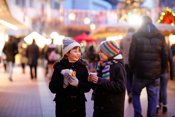 Cute little kids girl and boy having fun on traditional Christmas market during strong snowfall. Happy children eating traditional curry sausage called wurst and drinking hot chocolate. Twins friends — Stock Photo, Image