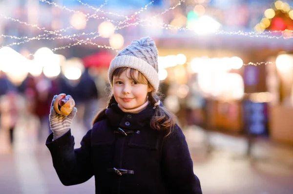 Roztomilé holčička baví na tradičním vánočním trhu během silného sněžení. Šťastné dítě jí tradiční kari klobásu zvanou Wurst. školačka stojící ozářený vánoční strom. — Stock fotografie