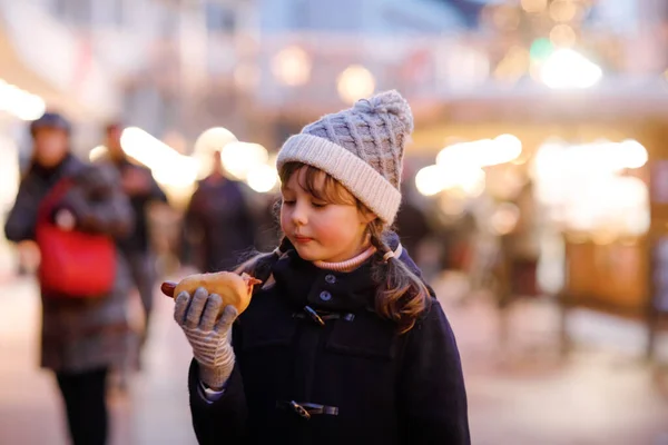 強い雪の中で伝統的なクリスマスマーケットで楽しんでかわいい子供の女の子。ウルトと呼ばれる伝統的なカレーソーセージを食べる幸せな子供。イルミネーションされたクリスマスツリーのそばに立つ女子高生. — ストック写真