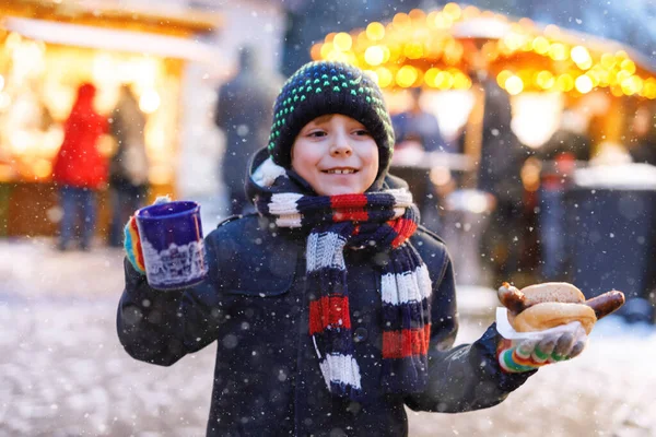 Pequeño niño lindo comiendo salchicha alemana y beber ponche niños calientes en el mercado de Navidad. Niño feliz en el mercado familiar tradicional en Alemania. Chico risueño en ropa de invierno colorida — Foto de Stock