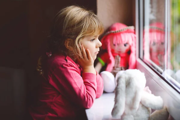 Petite fille mignonne assise à côté de la fenêtre et regardant dehors le jour de pluie. Enfant rêveur avec poupée et jouet doux se sentant heureux. Self isolation concept during corona virus pandemic time. Enfant solitaire. — Photo