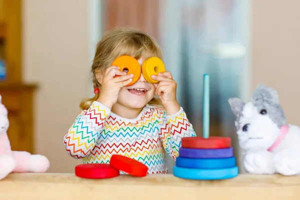 Linda niña pequeña jugando sola con colorida pirámide de arco iris de madera y juguetes en casa o guardería. Feliz niño sano que se divierte en el jardín de infantes o guardería preescolar —  Fotos de Stock