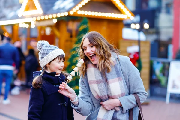 Mother and daughter eating white chocolate covered fruits and strawberry on skewer on traditional German Christmas market. Happy girl and woman on traditional family market in Germany during snowy day — Stock Photo, Image