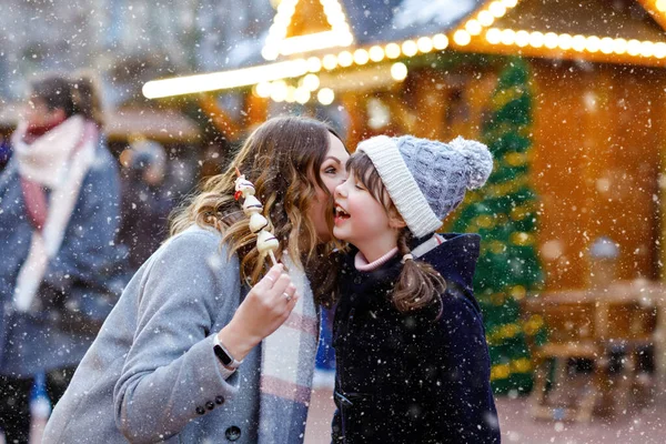 Mother and daughter eating white chocolate covered fruits and strawberry on skewer on traditional German Christmas market. Happy girl and woman on traditional family market in Germany during snowy day — Stock Photo, Image