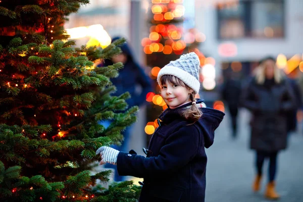 Kleine süße Mädchen, die sich bei starkem Schneefall auf dem traditionellen Weihnachtsmarkt vergnügen. Glückliches Kind beim traditionellen Familienmarkt in Deutschland. Schulmädchen steht am beleuchteten Weihnachtsbaum. — Stockfoto