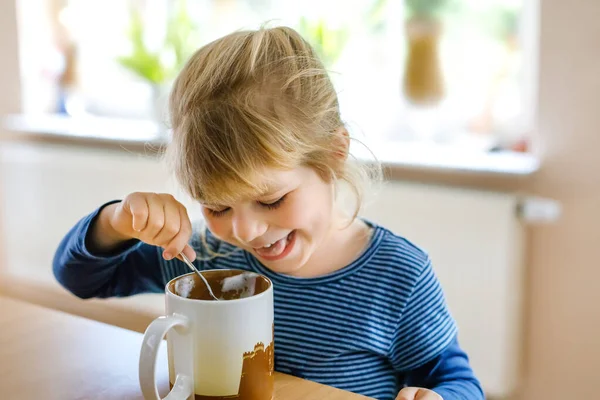愛らしい幼児の女の子は熱い牛乳を泡で飲んでいます。幸せな健康な子供室内、チョコレートドリンクを楽しんで. — ストック写真