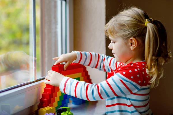 Roztomilé batole holčička po okně vytvořit duhu s barevnými plastovými bloky během pandemické koronavirus karantény. Děti vyrobeny a malovat duhy po celém světě jako znamení. — Stock fotografie