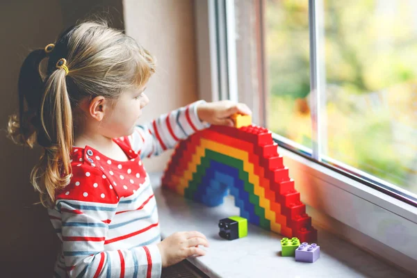 Linda niña pequeña por ventana crear arco iris con bloques de plástico de colores durante la cuarentena pandémica coronavirus. Los niños hicieron y pintaron arcoíris alrededor del mundo como signo. — Foto de Stock