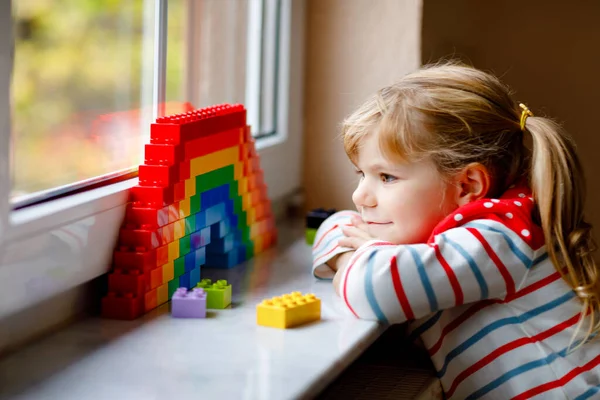 Schattig klein peuter meisje door het raam te creëren regenboog met kleurrijke plastic blokken tijdens pandemische coronavirus quarantaine. Kinderen maakten en schilderen regenbogen over de hele wereld als teken. — Stockfoto