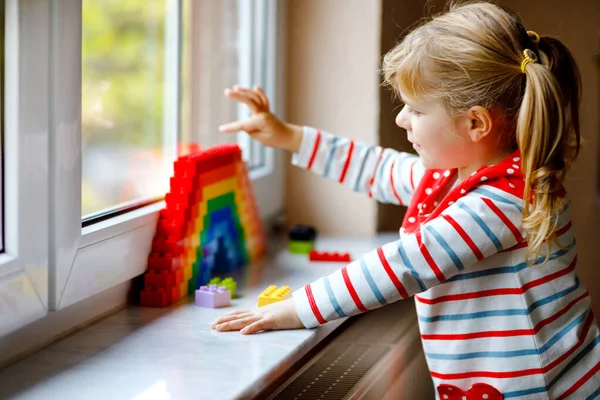 Linda niña pequeña por ventana crear arco iris con bloques de plástico de colores durante la cuarentena pandémica coronavirus. Los niños hicieron y pintaron arcoíris alrededor del mundo como signo. — Foto de Stock