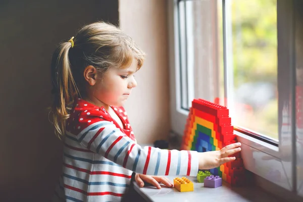 Linda niña pequeña por ventana crear arco iris con bloques de plástico de colores durante la cuarentena pandémica coronavirus. Los niños hicieron y pintaron arcoíris alrededor del mundo como signo. — Foto de Stock