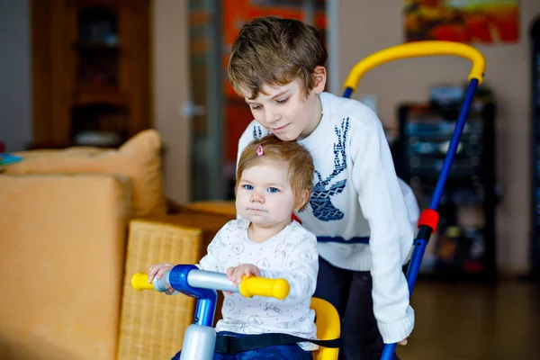 Garotinho empurrando bicicleta ou triciclo com a linda irmãzinha. Menina e irmão criança brincando juntos em casa, dentro de casa. Ligação de irmãos e amor, atividade conjunta e comum e jogo. — Fotografia de Stock