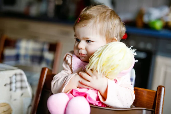 Bayi perempuan manis yang menggemaskan bermain dengan boneka pertama. Anak balita yang cantik duduk di kursi tinggi di rumah. Anak yang sehat bermain sendirian.. — Stok Foto