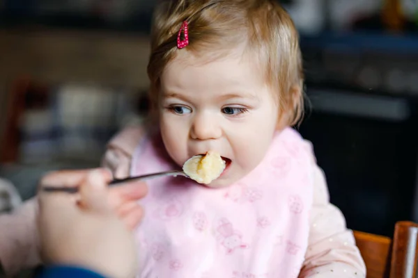 Entzückendes kleines Mädchen, das aus einem Löffel püriertem Gemüse und Püree isst. Essen, Kind, Ernährung und Menschen-Konzept -niedliches Kleinkind, Tochter mit Löffel sitzt im Hochstuhl und isst zu Hause. — Stockfoto