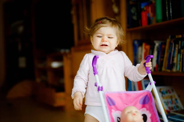 Cute adorable baby girl making first steps with doll carriage. Beautiful toddler child pushing stroller with toy at home. Happy daughter learning walking and standing