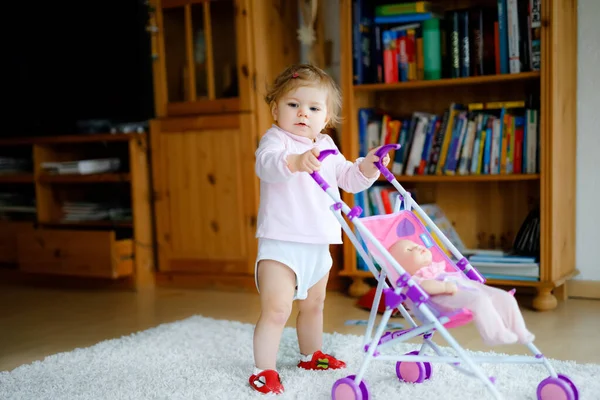 Симпатичная симпатичная девочка делает первые шаги с коляской. Красивый малыш толкает коляску с игрушкой дома. Счастливая дочь учится ходить и стоять — стоковое фото