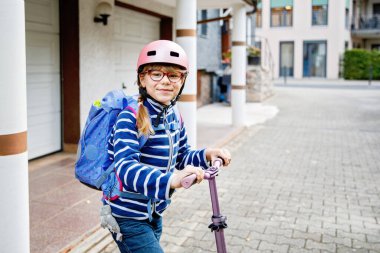 Okula kask ve sırt çantası takıp scooter süren mutlu ilkokul kızı. Güvenli, aktif, gerçek hayatta şehirde seyahat eden bir çocuk.
