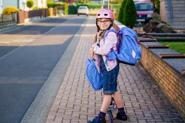 Okula kask ve sırt çantası takıp scooter süren mutlu ilkokul kızı. Güvenli, aktif, gerçek hayatta şehirde gezen sevimli bir çocuk.
