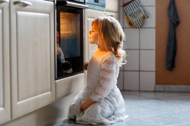 Şirin sarışın kız ev mutfağında kek ya da turta pişiriyor. Mutlu çocuk yardım ederken eğleniyor, Ofen 'in yanında oturuyor ve kurabiyeleri bekliyor. Çocuk Şükran Günü ya da Noel tatili için yemek pişiriyor.