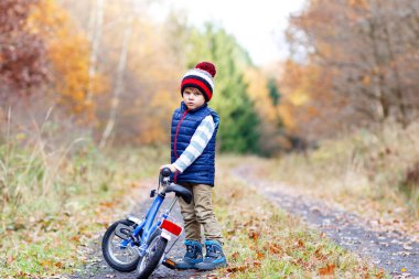 Sonbahar Ormanı Parkı 'nda renkli, sıcak giysiler içinde bisiklet süren küçük çocuk. Doğada güneşli bir sonbahar gününde bisiklet süren aktif bir çocuk. Güvenlik, spor, çocuk konseptiyle eğlence. Çocuklar için gündelik moda.