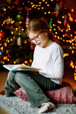Küçük liseli kız yılbaşı ağacının yanında ışıklarla kitap okuyor. Mutlu çocuk, aile tatili. Okul çocuğu kitap okuyor.