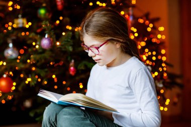 Küçük liseli kız yılbaşı ağacının yanında ışıklarla kitap okuyor. Mutlu çocuk, aile tatili. Okul çocuğu kitap okuyor.