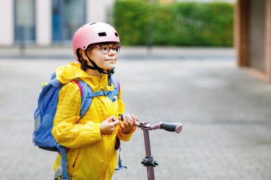 Okula kask ve sırt çantası takıp scooter süren mutlu ilkokul kızı. Güvenli, aktif, gerçek hayatta şehirde gezen sevimli bir çocuk.