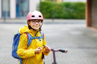 Okula kask ve sırt çantası takıp scooter süren mutlu ilkokul kızı. Güvenli, aktif, gerçek hayatta şehirde gezen sevimli bir çocuk.