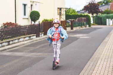 Okula kask ve sırt çantası takıp scooter süren mutlu ilkokul kızı. Güvenli, aktif, gerçek hayatta şehirde gezen sevimli bir çocuk.