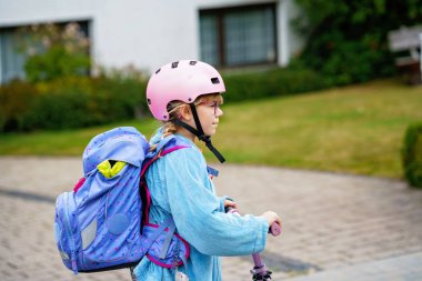 Okula kask ve sırt çantası takıp scooter süren mutlu ilkokul kızı. Güvenli, aktif, gerçek hayatta şehirde gezen sevimli bir çocuk.