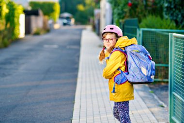 Okula kask ve sırt çantası takıp scooter süren mutlu ilkokul kızı. Güvenli, aktif, gerçek hayatta şehirde gezen sevimli bir çocuk.