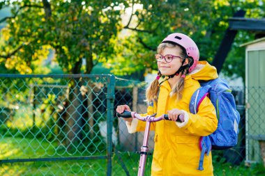 Okula kask ve sırt çantası takıp scooter süren mutlu ilkokul kızı. Güvenli, aktif, gerçek hayatta şehirde gezen sevimli bir çocuk.