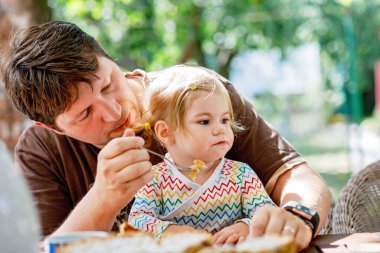 Sevimli küçük yürümeye başlayan çocuk kız restoranda besleme genç orta yaşlı baba. Arka plan üzerinde yemek çocuk evlat. Yaz saati, açık bir kafe mutlu sağlıklı aile pasta yemeye.