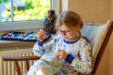 Küçük kız doldurulmuş hayvanı için yastık dikiyor. Mutlu Çocuk elinde iğne iplikle pencerenin yanında oturuyor..