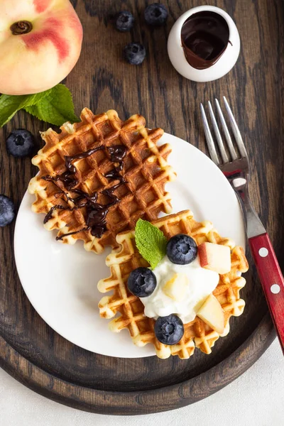 Belgian waffles with fresh blueberries, peaches and chocolate sauce on a rustic wooden cutting board. Liege style belgian waffles. Breakfast or lunch concept.