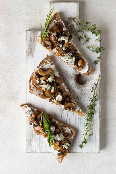 Whole grain rye bread toast with cream cheese or ricotta, mushroom and herbs on a white wooden cutting board.