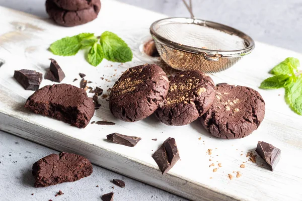 Chocolate cookies, pieces of chocolate, mint and cocoa powder on a white wooden cutting board.