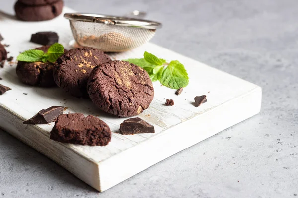 Chocolate cookies, pieces of chocolate, mint and cocoa powder on a white wooden cutting board.