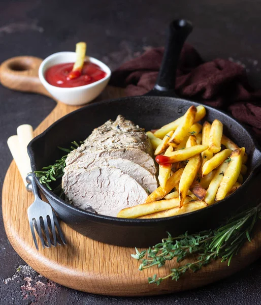 A skillet with roast pork meat served with potato fries and herbs on brown stone table.