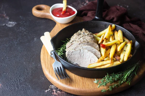 A skillet with roast pork meat served with potato fries and herbs on brown stone table.